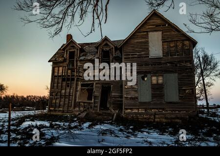 A wide angle horizontal shot of a creepy, abandoned old wooden house with windows rotting and wood falling apart, and snow outside at sunset in the wi Stock Photo