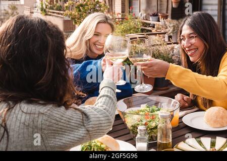 Multiracial friends eating vegan food cheering with wine outdoors at patio restaurant - Focus on center girl eye Stock Photo