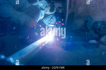 Welder welding metal with argon arc welding machine and has welding sparks. A man wears welding mask and protective gloves. Safety in industrial Stock Photo