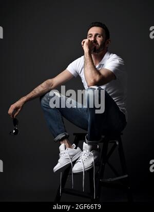 Thoughtful unshaved man in white polo shirt, modern torn jeans and sneakers sits on high chair, stepladder Stock Photo