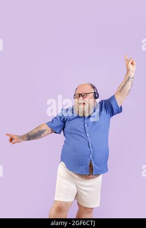 Positive man with overweight enjoys favourite music dancing on purple background Stock Photo