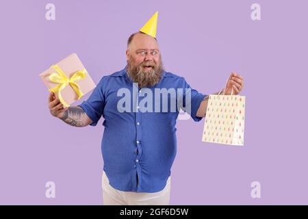 Excited fat man in party hat holds gift and paper bag on purple background Stock Photo