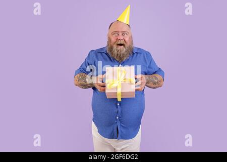 Excited plump man wearing tight shirt holds present on purple background Stock Photo