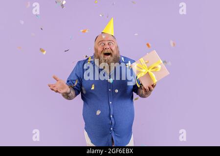 Emotional plus size man holds gift box standing under confetti shower on purple background Stock Photo