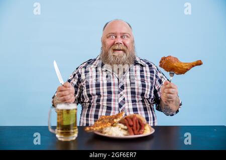 Smiling man with overweight holds chicken leg sitting at table on light blue background Stock Photo