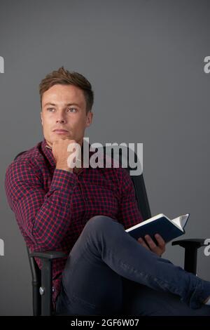 Portrait of pensive handsome young man sitting in chair and filling gratitude journal or writing down ideas Stock Photo
