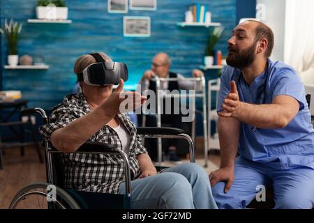 Male nurse giving social support, helping retired disabled senior woman to enjoy virtual reality using headset during caregiver service. Social nursing services at home. Healthcare assistance Stock Photo