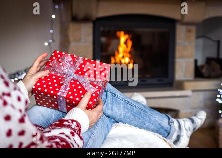 Woman holding christmas gift box in hands near fireplace in cozy home interior. Woman sitting at chair and opening christmas present Stock Photo