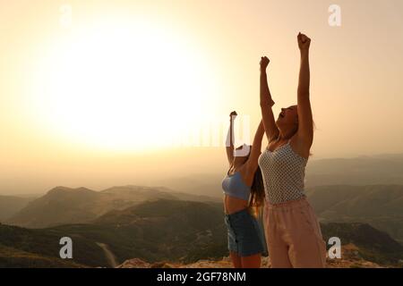 Two happy friends raising arms celebrating vacation at sunset in the mountain