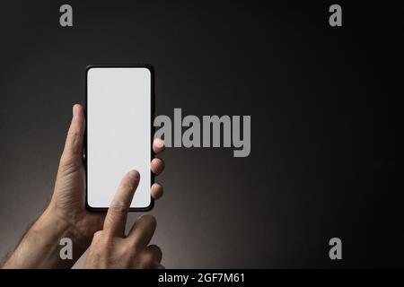 Colour image of man's hand holding black smartphone using blank white screen with finger, on dark background, dramatic lighting and copy space to side Stock Photo