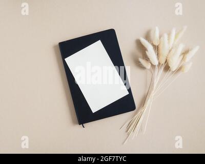 White blank card for text on black nptepad next to bouquet of dry Lagurus on beige background. Minimal concept for message, message or invitation in p Stock Photo