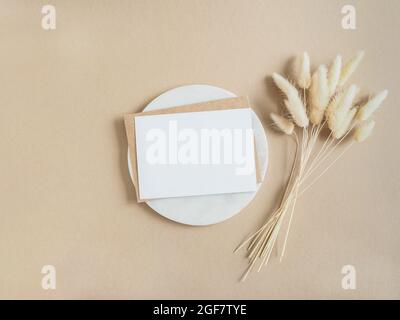 White blank card for text and envelope on marble round podium next to bouquet of dry Lagurus on beige background. Minimal concept for message, message Stock Photo