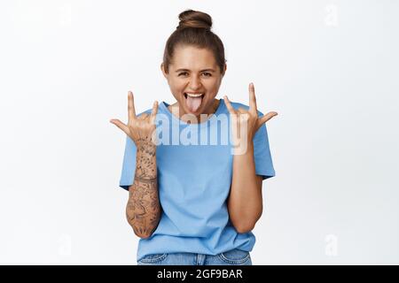Joyful girl having fun, showing rock on heavy metal horns, sticking tongue and shouting excited, standing in blue t-shirt against white background Stock Photo