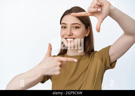 Snap. Smiling happy girl taking picture, looking through hand frame gesture as if photographing with camera, standing in tshirt against white Stock Photo