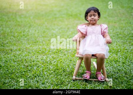Cute little Asian girl with closed eyes sitting on a chair on the lawn Stock Photo