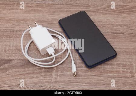 On a wooden table there is a cell phone and a charger with its USB cable. The cable is white and coiled. The smartphone is turned off. Stock Photo
