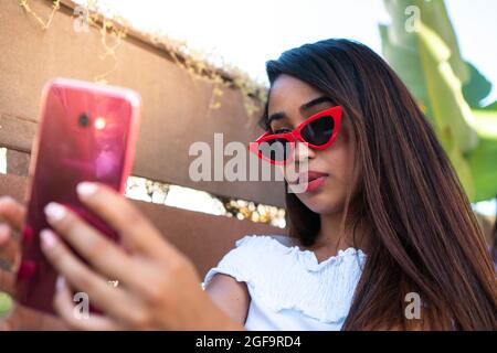 Latin woman taking a selfie. Hispanic girl taking a phone of herself with red glasses. Beautiful brunette woman looking at the phone with a serious fa Stock Photo