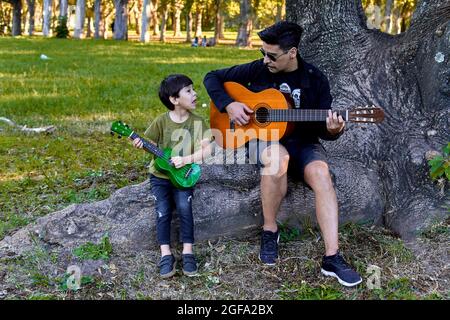 Latin father and son playing guitar and ukelele together sitting i a tree in a forest in Argentina. Horizontal, blurred background Stock Photo
