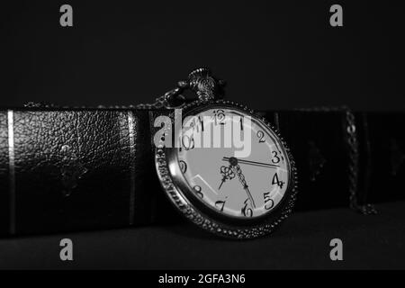 Old pocket watch resting against black leather book, photographed in black and white Stock Photo