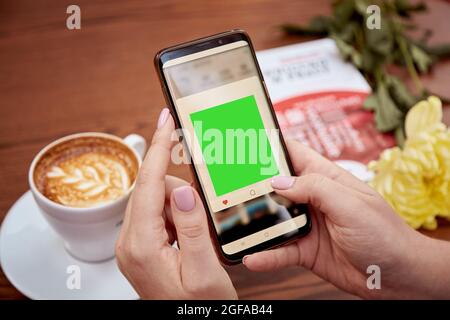 Chroma key mobile phone screen in woman's hand and cup of coffee on the table. Top view. Education, working, freelance concept. High quality photo Stock Photo