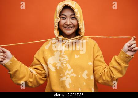 Asian teen girl wearing hoodie smiling and looking at camera isolated over orange background Stock Photo
