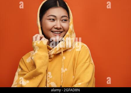 Asian teen girl wearing hoodie smiling and looking aside isolated over orange background Stock Photo