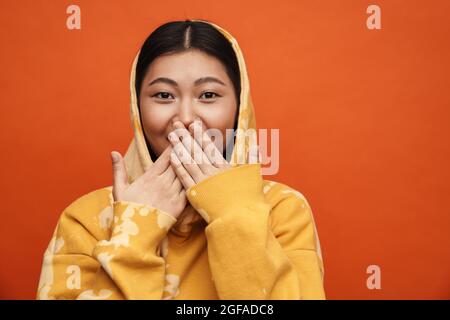 Asian teen girl wearing hoodie covering her mouth isolated over orange background Stock Photo