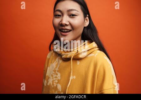 Asian teen girl wearing hoodie smiling and looking at camera isolated over orange background Stock Photo