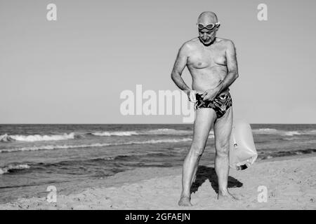 Sport swimmer senior man training to swim open water triathlon with beach on background - Elderly and healthy lifestyle - Black and white edit Stock Photo