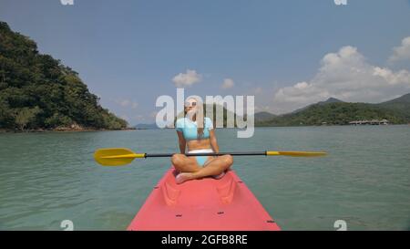 Attractive sportive blonde woman with sunglasses rows pink plastic canoe along sea water against green hills and blue sky. Traveling to tropical count Stock Photo