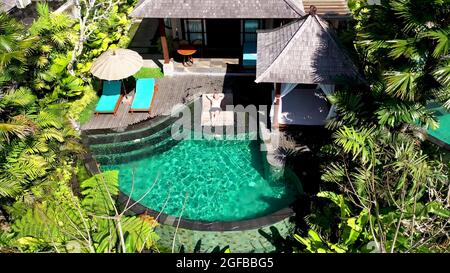 Aerial top view from drone, male tourist in black swimsuit lying on sun bed in contemporary villa surrounded by tropical palm trees. Property rental d