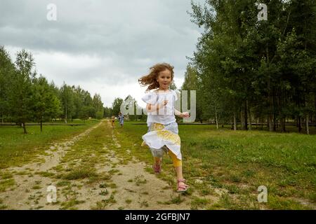 Funny girl in skirt and blouse running on road towards camera against cloudy sky while spending weekend day near grove in nature Stock Photo