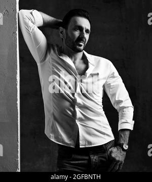 Black and white. Unshaved adult man in white shirt and jeans stands at concrete wall, leaning, holding hand behind head Stock Photo