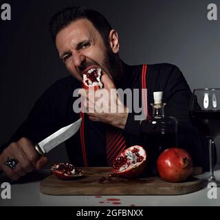Brunet man in black shirt, tie and suspenders sits at table with bottle and glass of fresh juice and bites pomegranate Stock Photo
