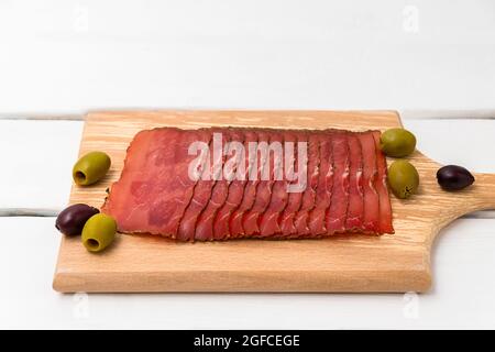 Fin slices of dried meat on cutting board with olives on white wooden table