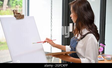 Blank canvas easel mockup for your artwork painting by attractive female artist, holding colour palette Stock Photo