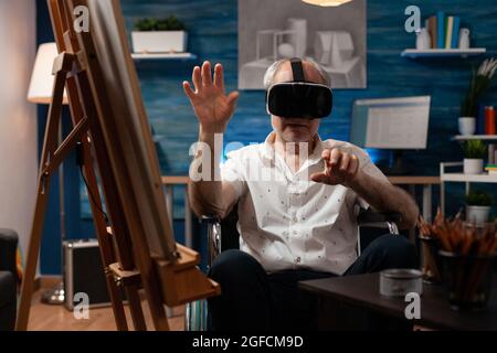 Invalid aged artist using vr glasses to draw masterpiece design on canvas and easel at fine art studio. Disabled senior man with modern headset sitting in wheelchair, for drawing hobby Stock Photo
