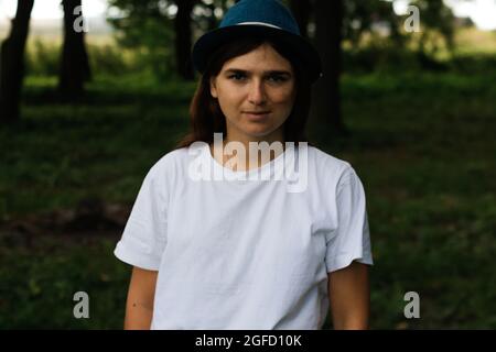 Defocus close-up portrait of a tired young woman with brown hair wearing a hat outdoors. Green nature background. Looking at camera. Women standing at Stock Photo