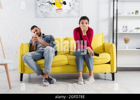 Scared african american woman holding remote controller near boyfriend using smartphone on couch Stock Photo