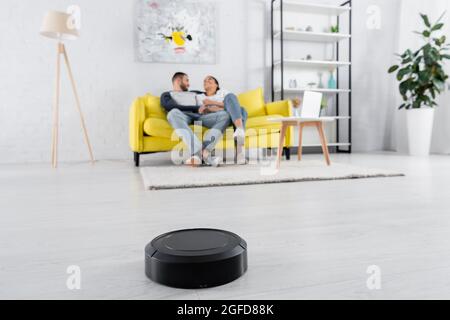 Robotic vacuum cleaner on floor near blurred couple in living room Stock Photo
