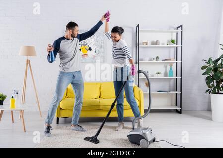 Happy multiethnic couple with cleaning supplies dancing in living room Stock Photo