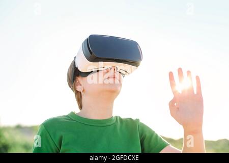 Young beautiful slim teenage girl wearing virtual reality glasses in outdoor nature background. Smartphone using with VR goggles headset in sunset Stock Photo