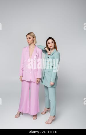 full length of blonde and redhead women in pink and blue pastel suits posing on grey Stock Photo