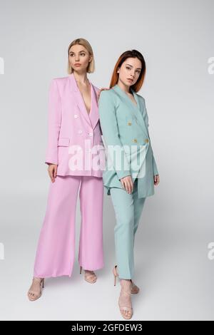 full length of young blonde and redhead models in pink and blue pastel suits posing on grey Stock Photo