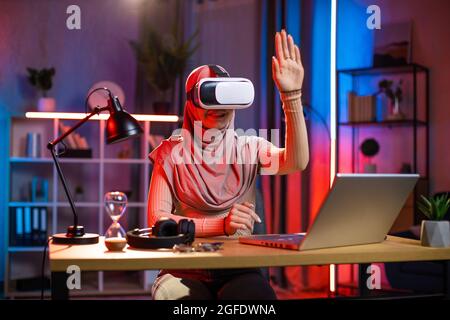 Young arabian woman sitting at home office and using VR headset and laptop for work during evening time. Concept of people, technologies and freelance. Stock Photo