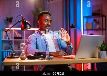 Young african american man waving and smiling during video call on modern laptop. Handsome guy sitting at home office and having online conference during evening time. Stock Photo