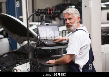 Elderly car mechanic smiling to the camera while repairing an auto at the workshop, using computer Stock Photo