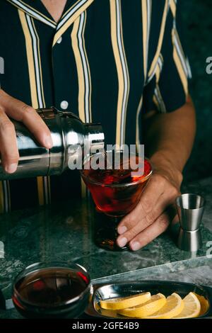 a young caucasian man, wearing a dark blue striped shirt, pours a red cocktail, from a metal shaker, in a glass placed on a green stone surface Stock Photo