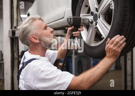 Bearded elderly car mecganic using electric torque wrench on car wheel Stock Photo