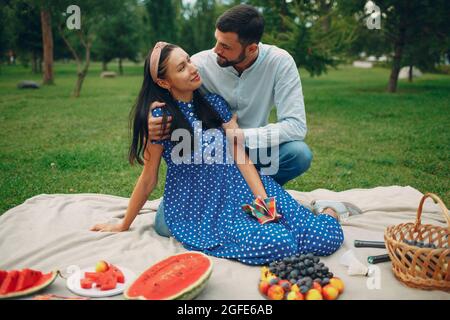 Young adult woman and man couple picnic at green grass meadow in park with fruits and basket Stock Photo